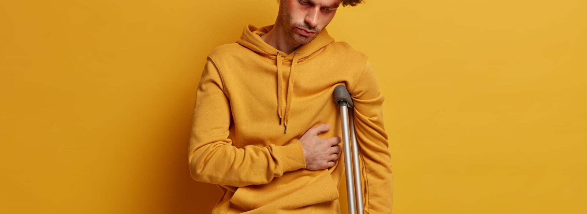 Indoor shot of distressed man has broken rib, suffers from pain, stands on crutches, had accident on