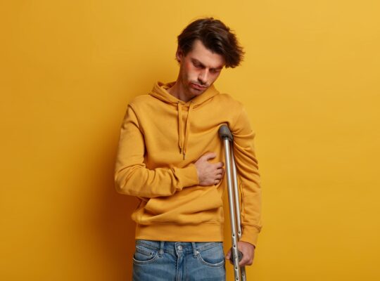Indoor shot of distressed man has broken rib, suffers from pain, stands on crutches, had accident on
