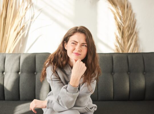 young woman sits on couch discomfort and squints. bored and sad woman with no mood