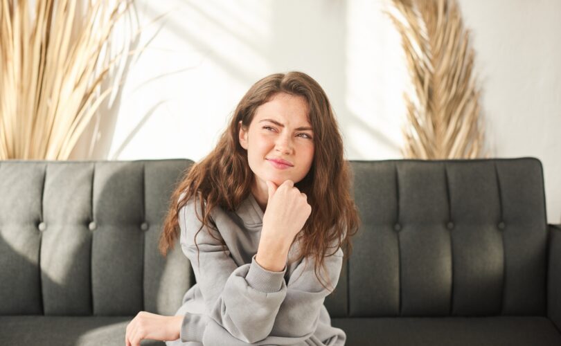 young woman sits on couch discomfort and squints. bored and sad woman with no mood